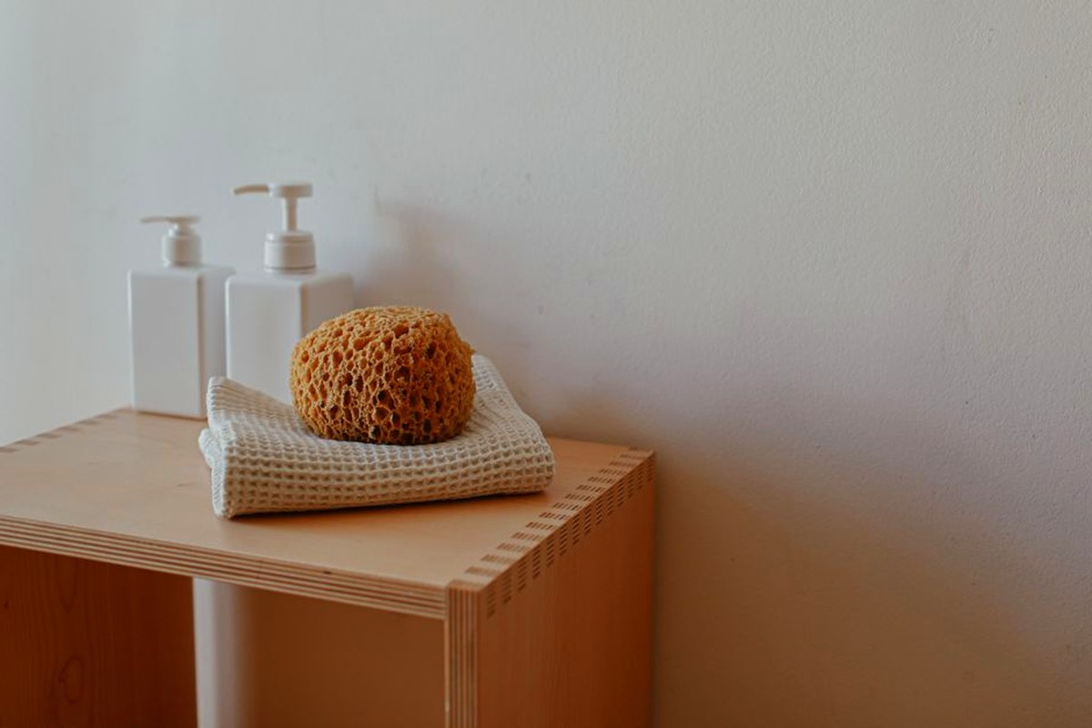 Close-up of a bathroom shelf featuring a natural sponge, towels, and pump bottles.